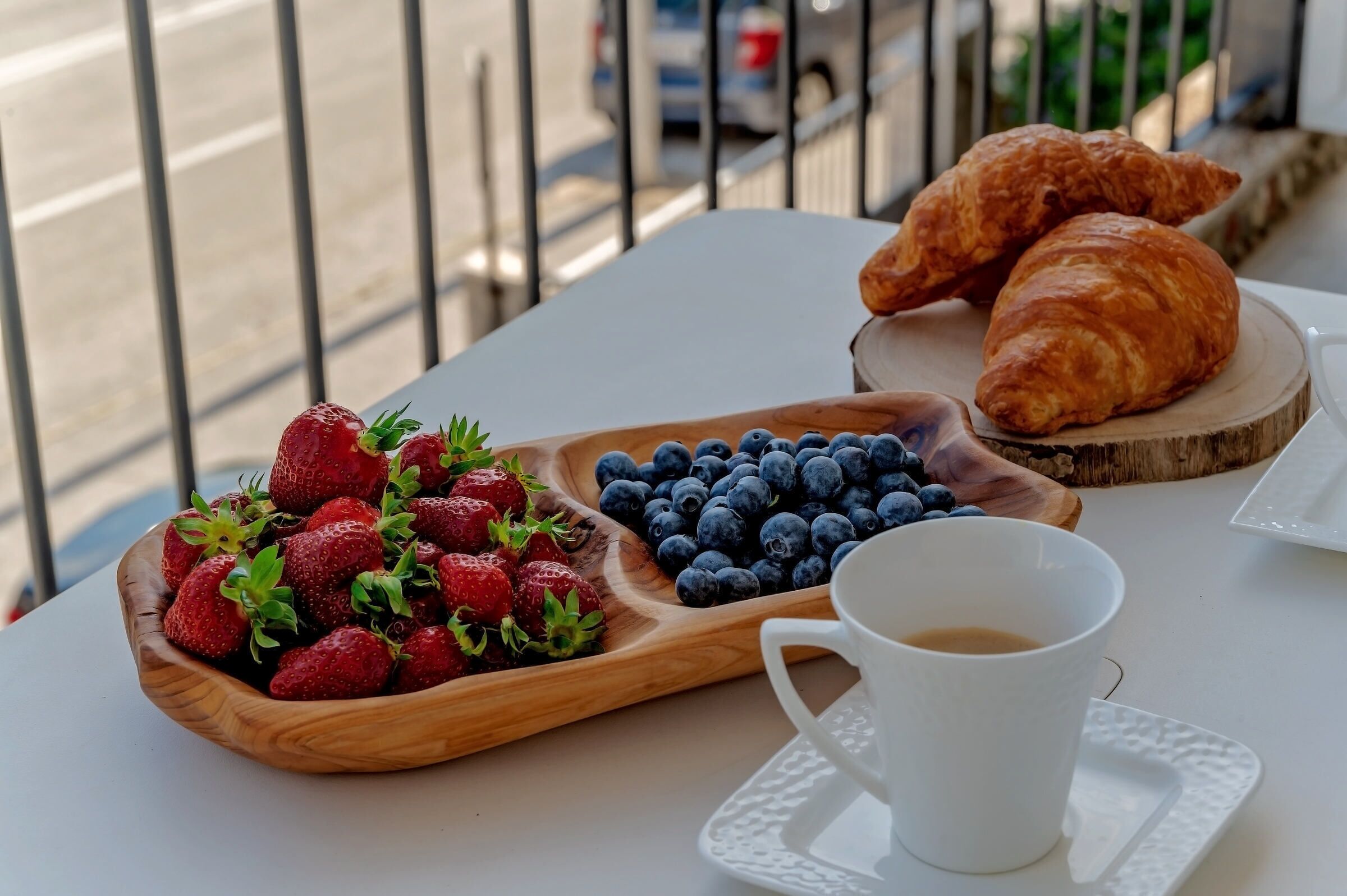 Una mesa con una tabla de fresas y arándanos, una taza de café y dos cruasanes sobre un mantel blanco.