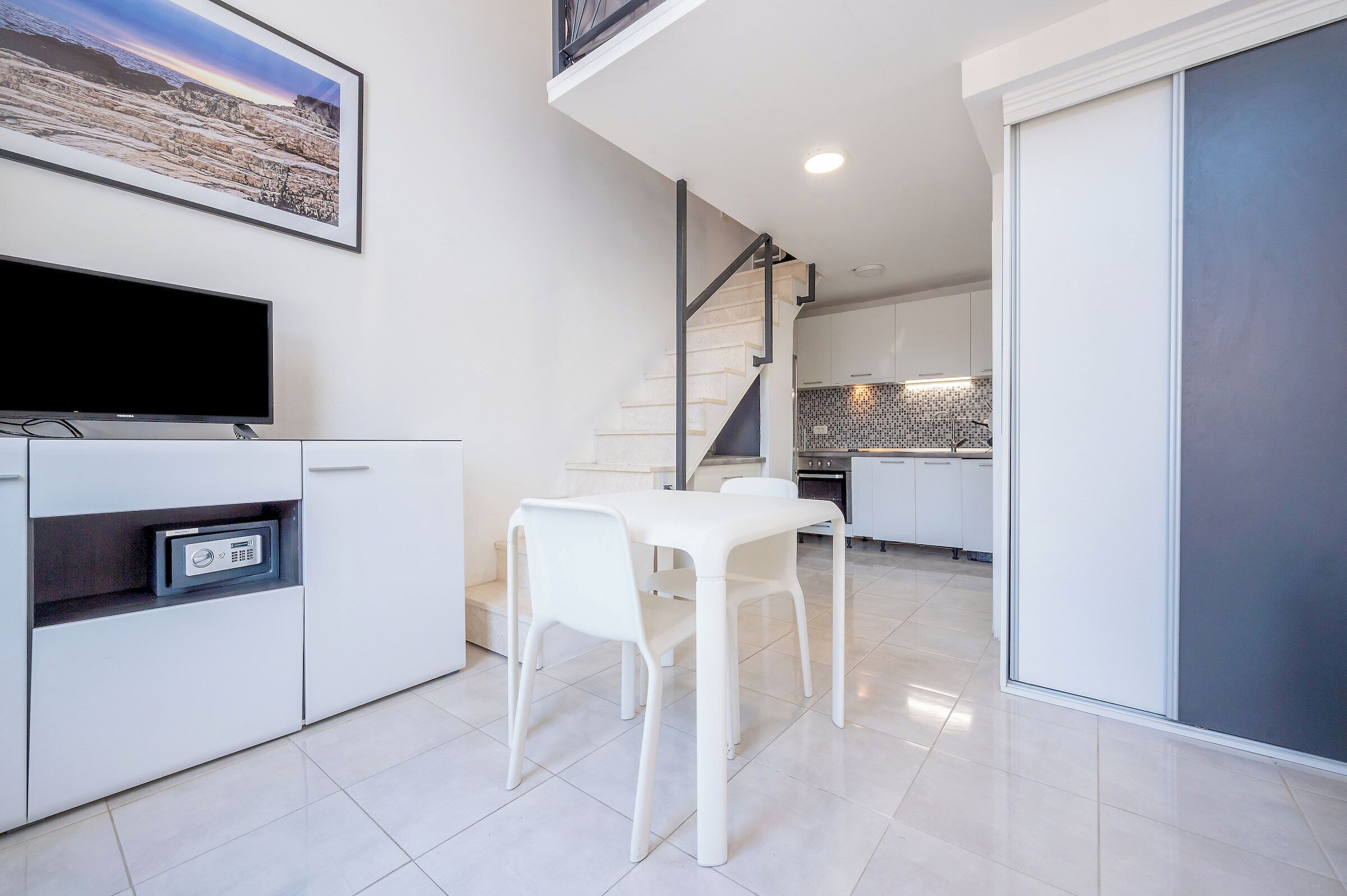 Una cuina moderna amb armaris blancs, una taula de menjador amb caires, un televisor de pantalla plana i una escala que mena a un nivell superior.