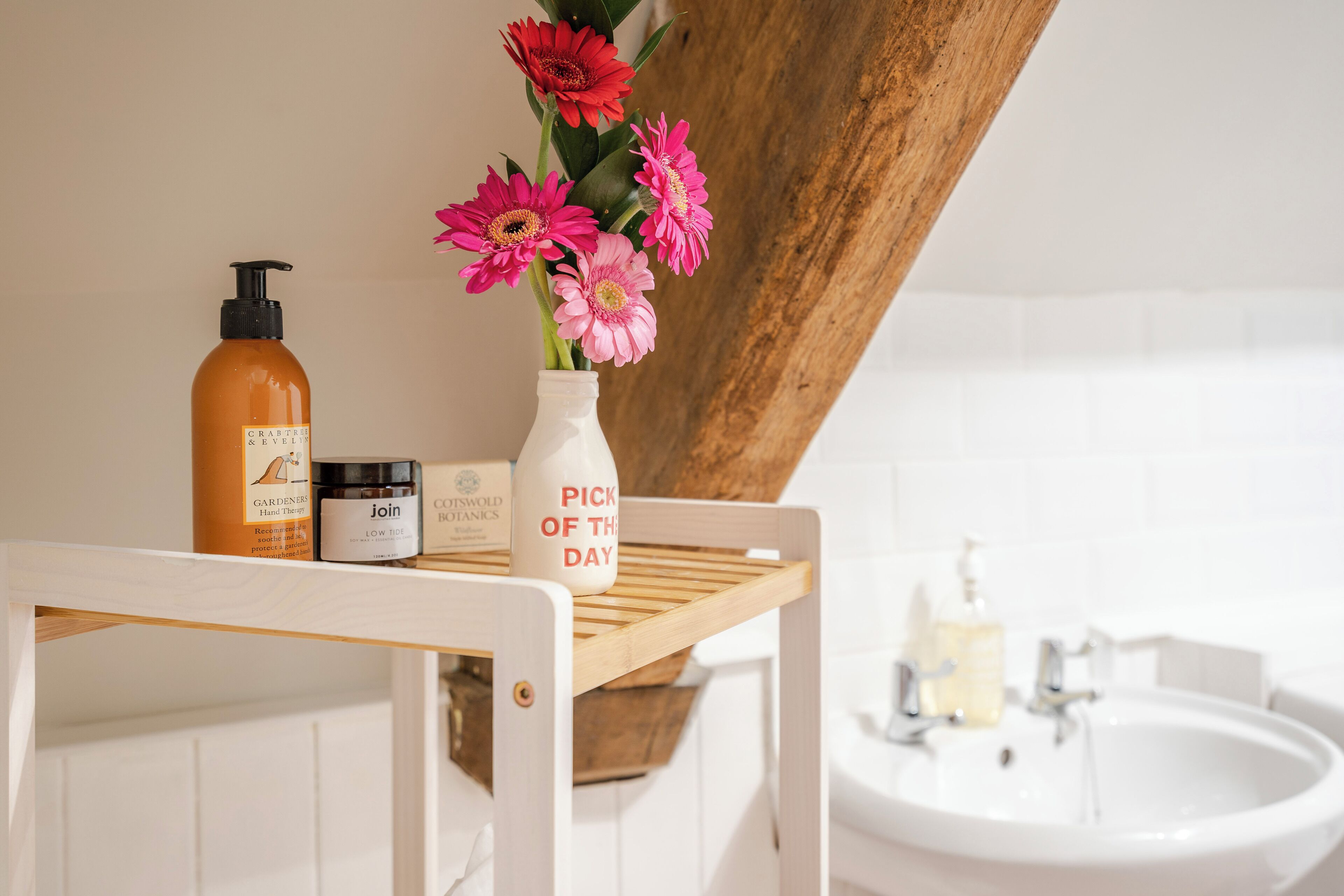 A bathroom vanity with a white sink, a wooden shelf holding toiletries, and a vase with pink flowers.