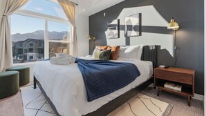 Ein modernes Schlafzimmer mit einem großen Bett, einem Nachttisch, einer Stehlampe und Bergblick durch ein großes Fenster.