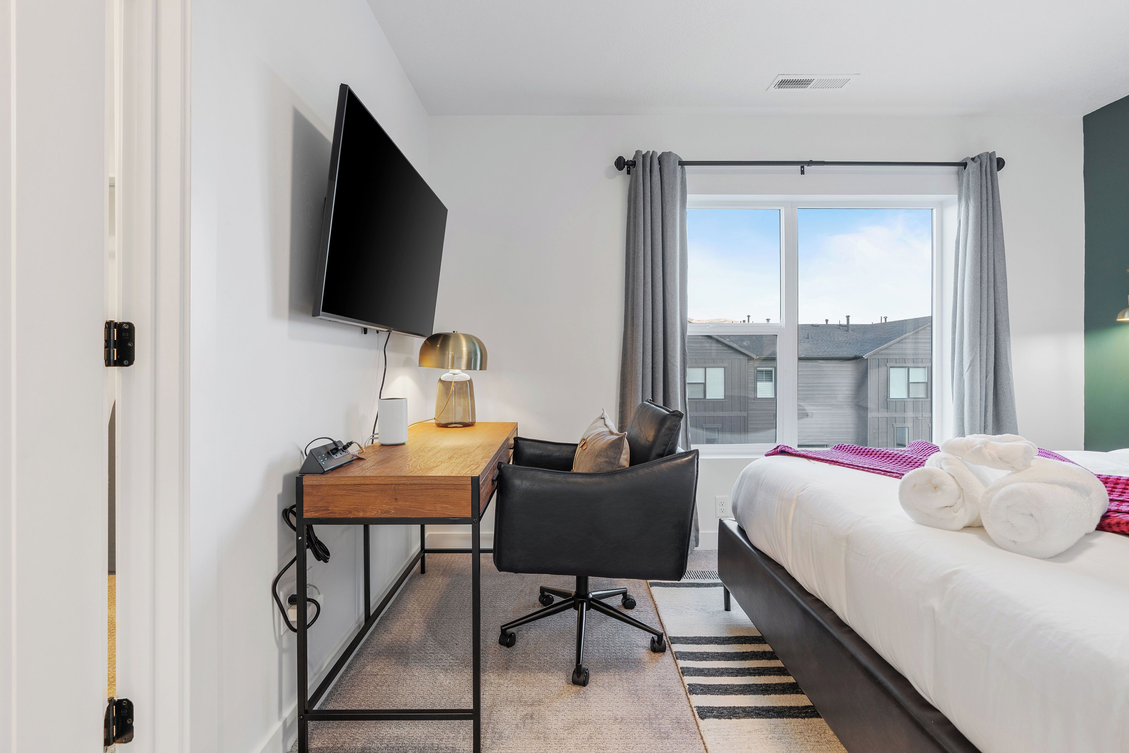 A hotel room with a bed, a desk with a chair, a TV mounted on the wall, and a window with curtains.