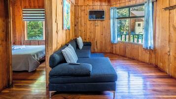 Una cabaña acogedora con un sofá azul oscuro, una cama con sábana blanca, paredes de madera y una ventana con vista a la vegetación.