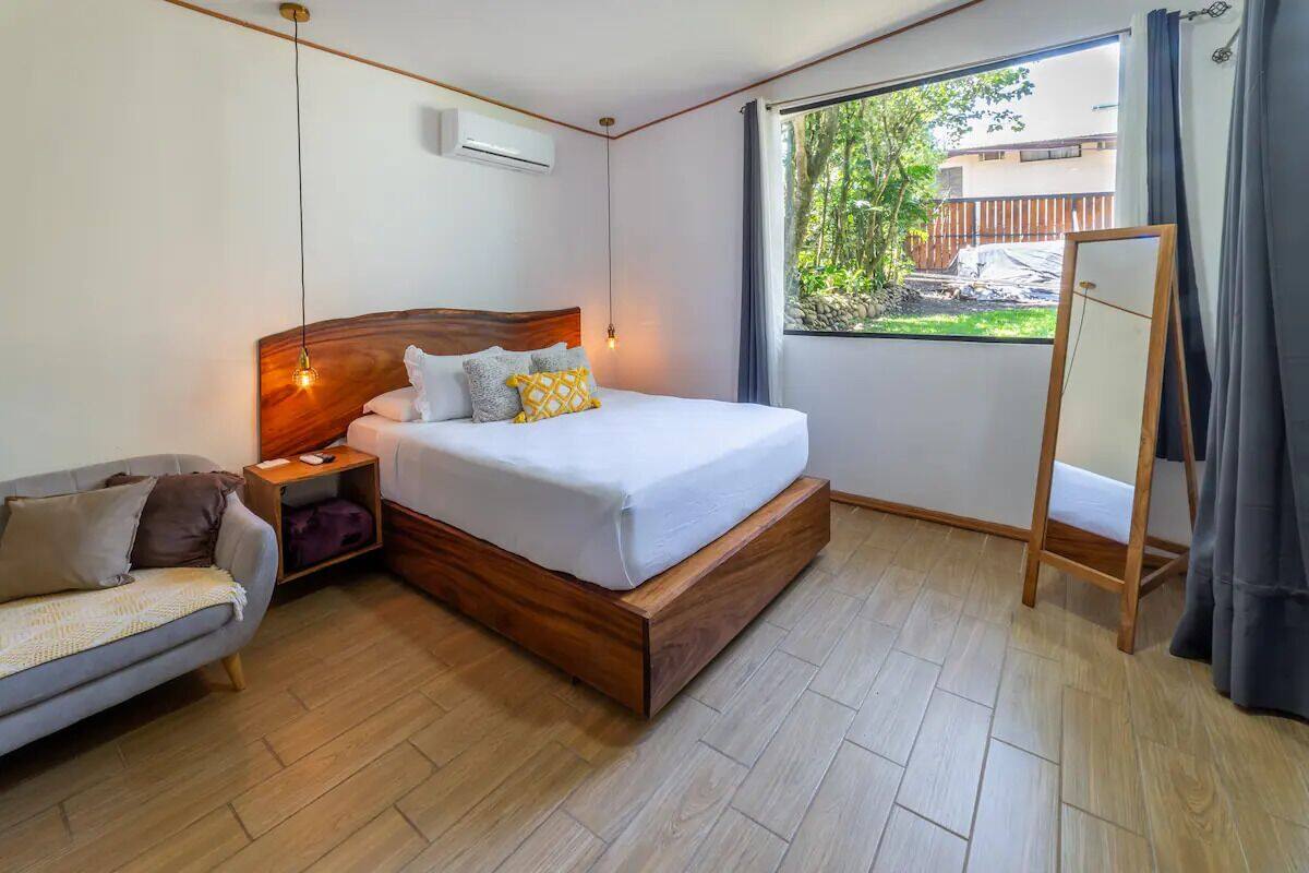 Ein Schlafzimmer mit einem hölzernen Bett, einem Nachttisch, einem Sofa, einem Fenster mit Blick auf Grünflächen und einem stehenden Spiegel.