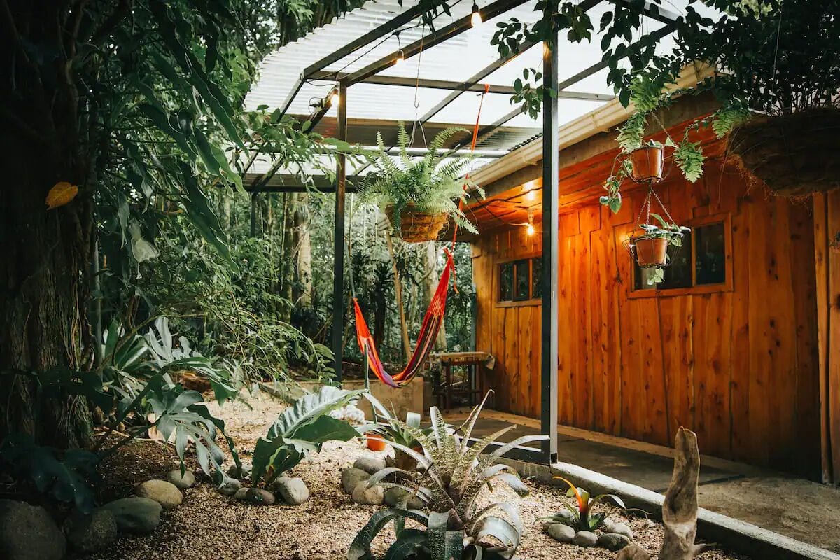 Eine Holzhütte mit einer Veranda, umgeben von üppiger Vegetation und hängenden Pflanzen.