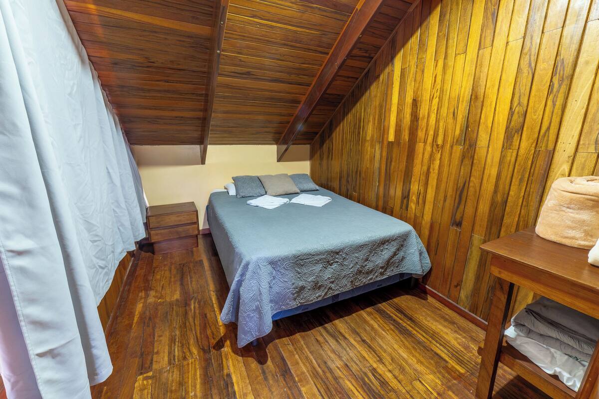 Ein kleiner, gemütlicher Raum mit Holzbalkendecke und -wänden, einem Einzelbett mit blauer Bettdecke, einem Nachttisch und einem Holzregal mit gefalteten Handtüchern.