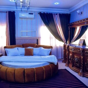A large, round bed with a brown headboard, a wooden dresser with a mirror, and a window with curtains. - Somewhere Apartments (Abuja)