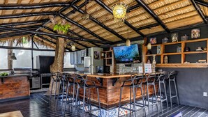 A bar area with a wooden counter and bar stools, a television, and a shelf with decorative items. - Villa Twelve27 (Sosúa)