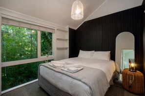 Une chambre d’hôtel moderne avec une grande fenêtre, un lit avec des serviettes soigneusement pliées, une lampe de chevet et un mur d’accent de couleur foncée.
