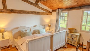Ein Schlafzimmer mit Holzbalkendecke, einem weißen Bett, einem Nachttisch mit Lampe, einer Kommode mit Schubladen und einem Fenster mit Vorhängen.