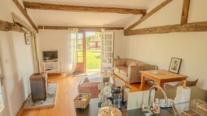 Ein gemütliches Zimmer mit Holzbalkendecke, einer Couch, einem Tisch, einem Fernseher und einer Küchenzeile mit Spüle sowie Blick auf den Garten.