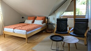 Une chambre avec un plafond en pente, un lit avec une literie orange, deux chaises noires, une petite table et une fenêtre avec des stores.