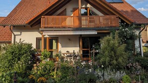 Une maison à deux étages avec un balcon, un jardin agrémenté de diverses plantes et un panneau solaire sur le toit.