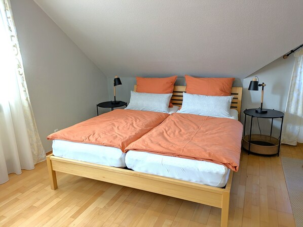 Ein ordentlich bezogenes Bett mit Holzbettgestell und einer pfirsichfarbenen Tagesdecke, zwei Nachttische mit Lampen und ein Korbwäschekorb auf dem Boden.