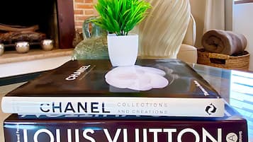 A living room with a TV displaying a movie, a fireplace, a lamp, a potted plant, and a stack of books including 'Chanel' and 'Louis Vuitton'.