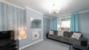 A modern living room with a flat-screen TV, a sofa set, a chandelier, and a large window with curtains.