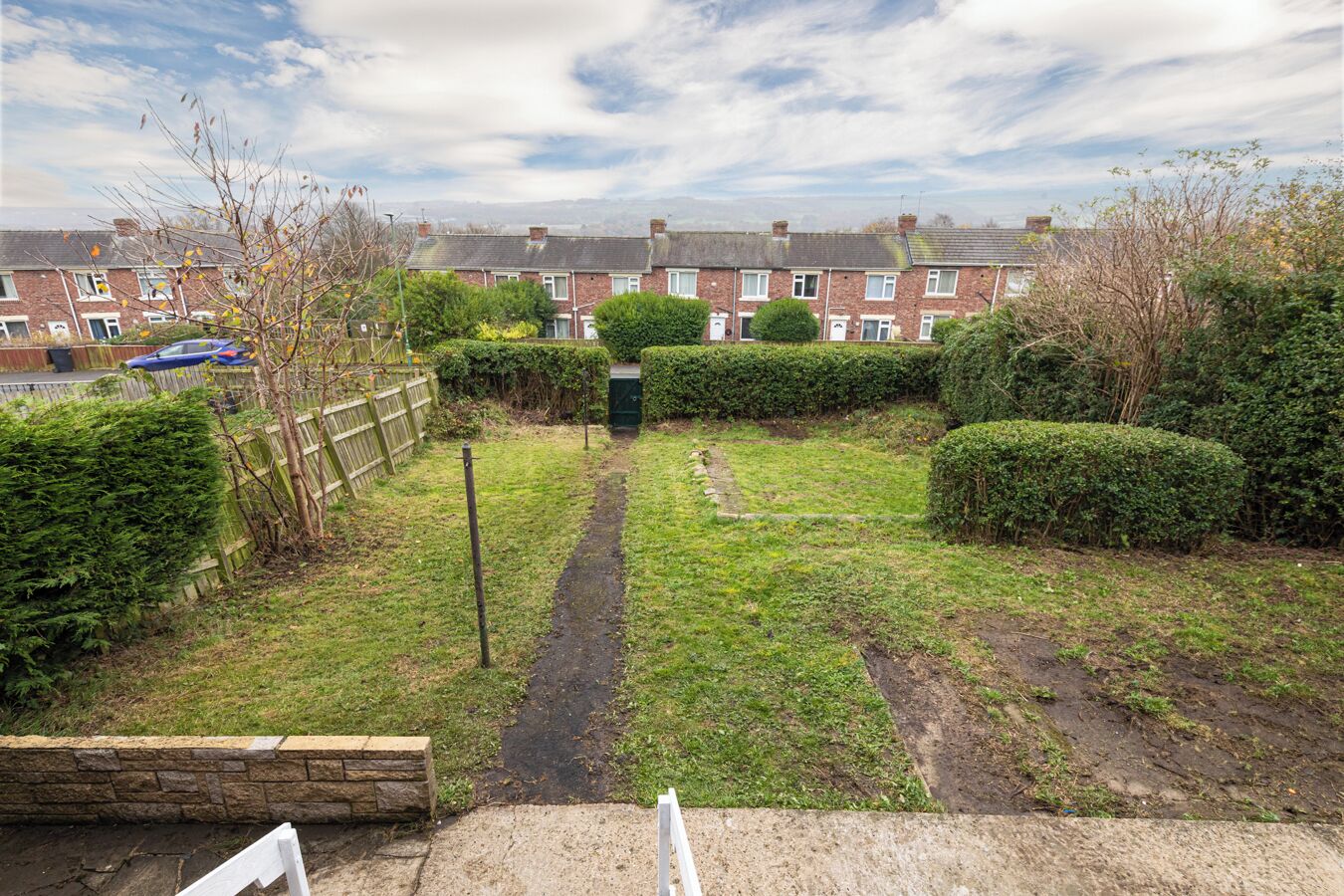 A residential garden with a paved path, hedges, and a brick house in the background.