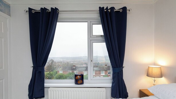 A bedroom with a bed, a window with curtains, a radiator, a lamp, and a view of the outside.