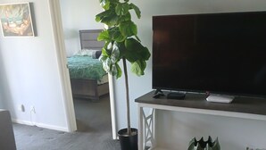 A living room with a TV on a white stand, a wooden table, a potted plant, and a bed in the background. - Naples Florida Paradise - 2 week Min Stays (Naples)