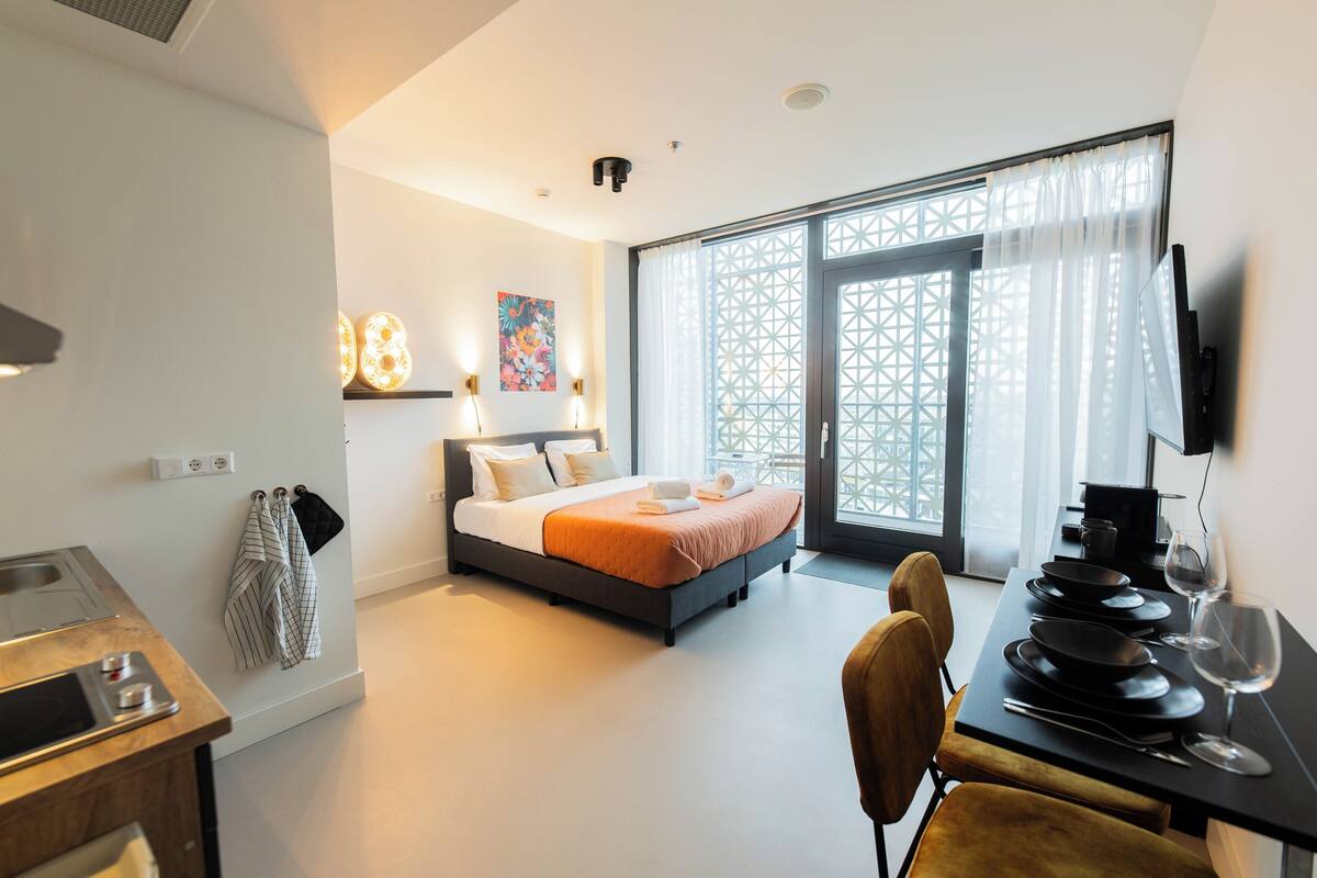 A modern hotel room with a bed, a dining table with plates and cutlery, a TV, and a large window with curtains.