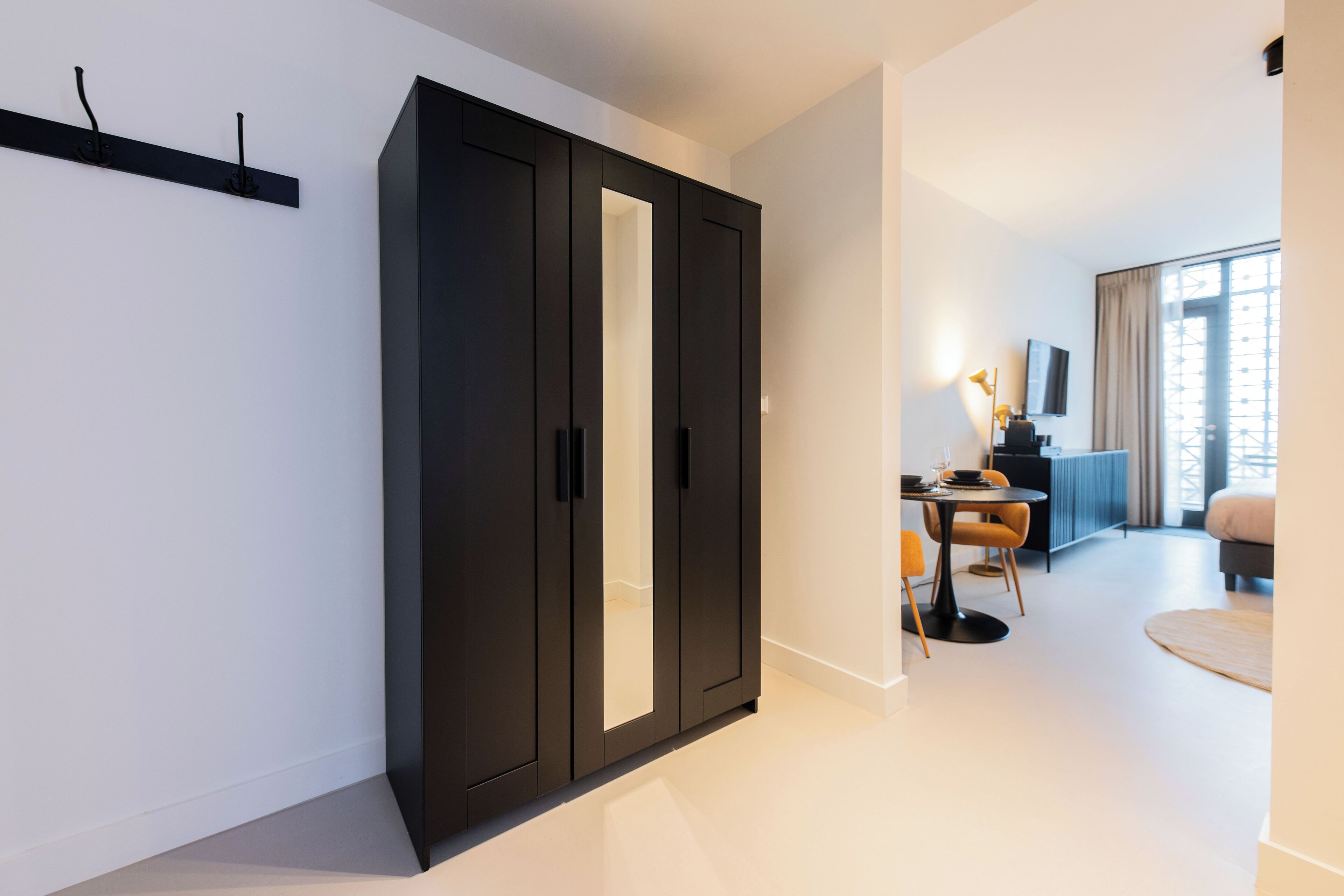 A modern hotel room with a large wardrobe, a dining area with a table and chairs, and a large window with curtains.