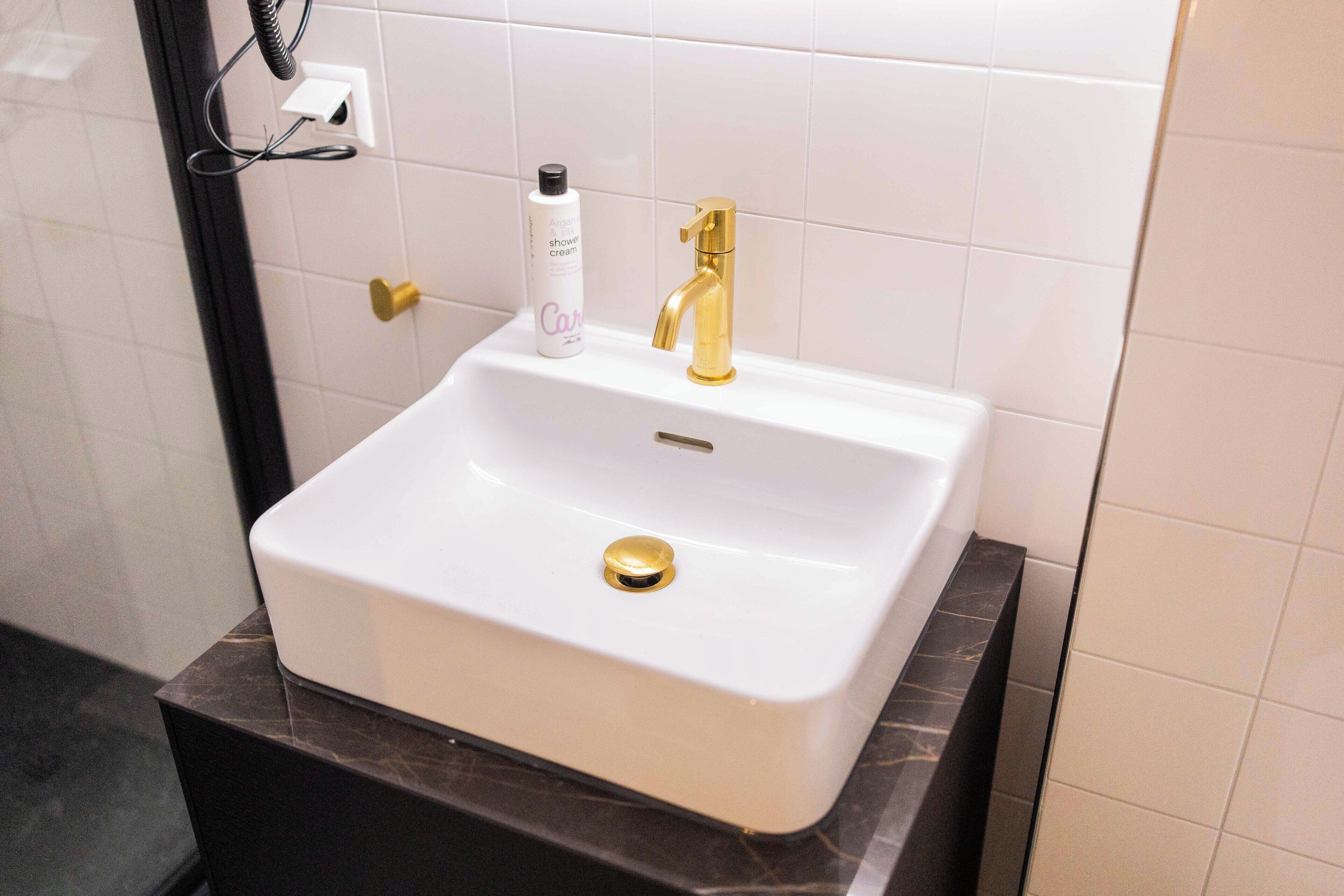 A bathroom with a white sink, gold faucet, and a bottle of shower cream.