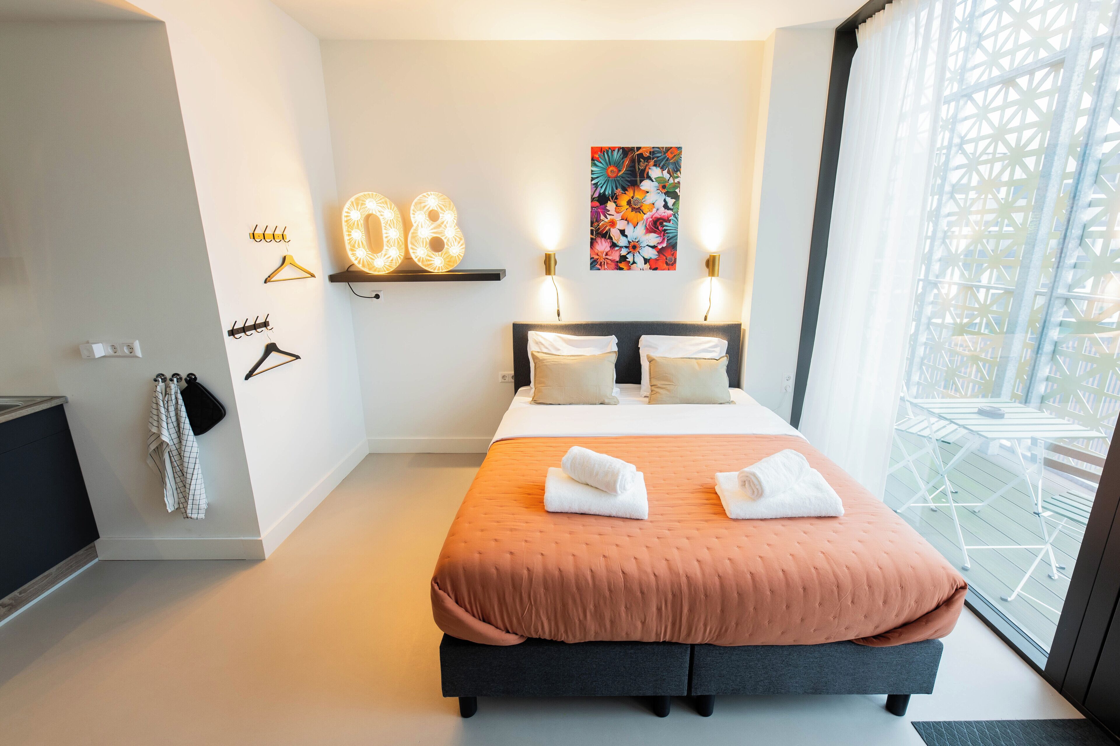 Une chambre moderne avec un grand lit, deux lampes de chevet, une étagère murale avec des luminaires et une œuvre d’art florale aux couleurs vives.
