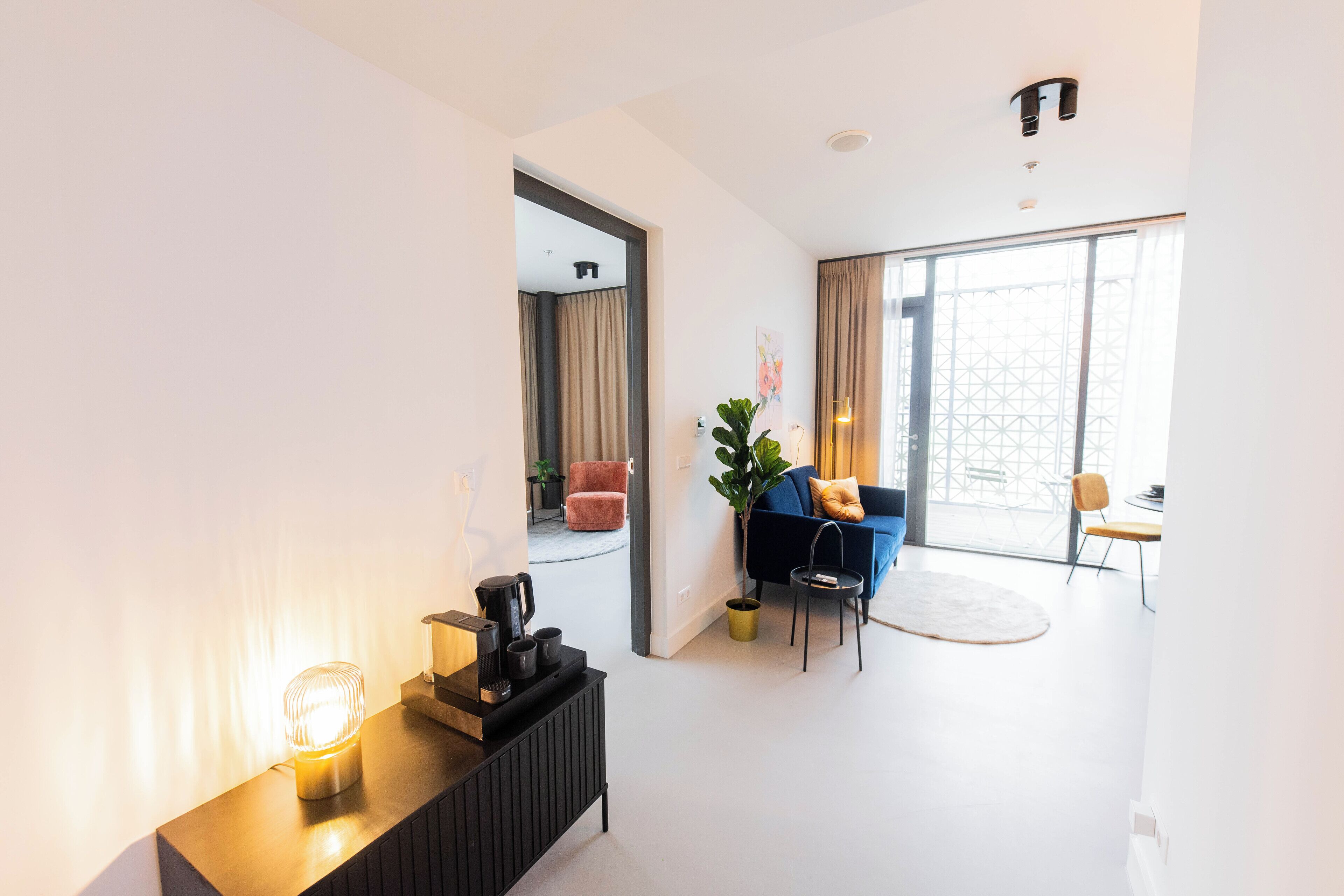 Een moderne woonkamer met een bank, een salontafel en een plant. Er is een groot raam met gordijnen, een deur die naar een andere kamer leidt, en een kleine tafel met een lamp.