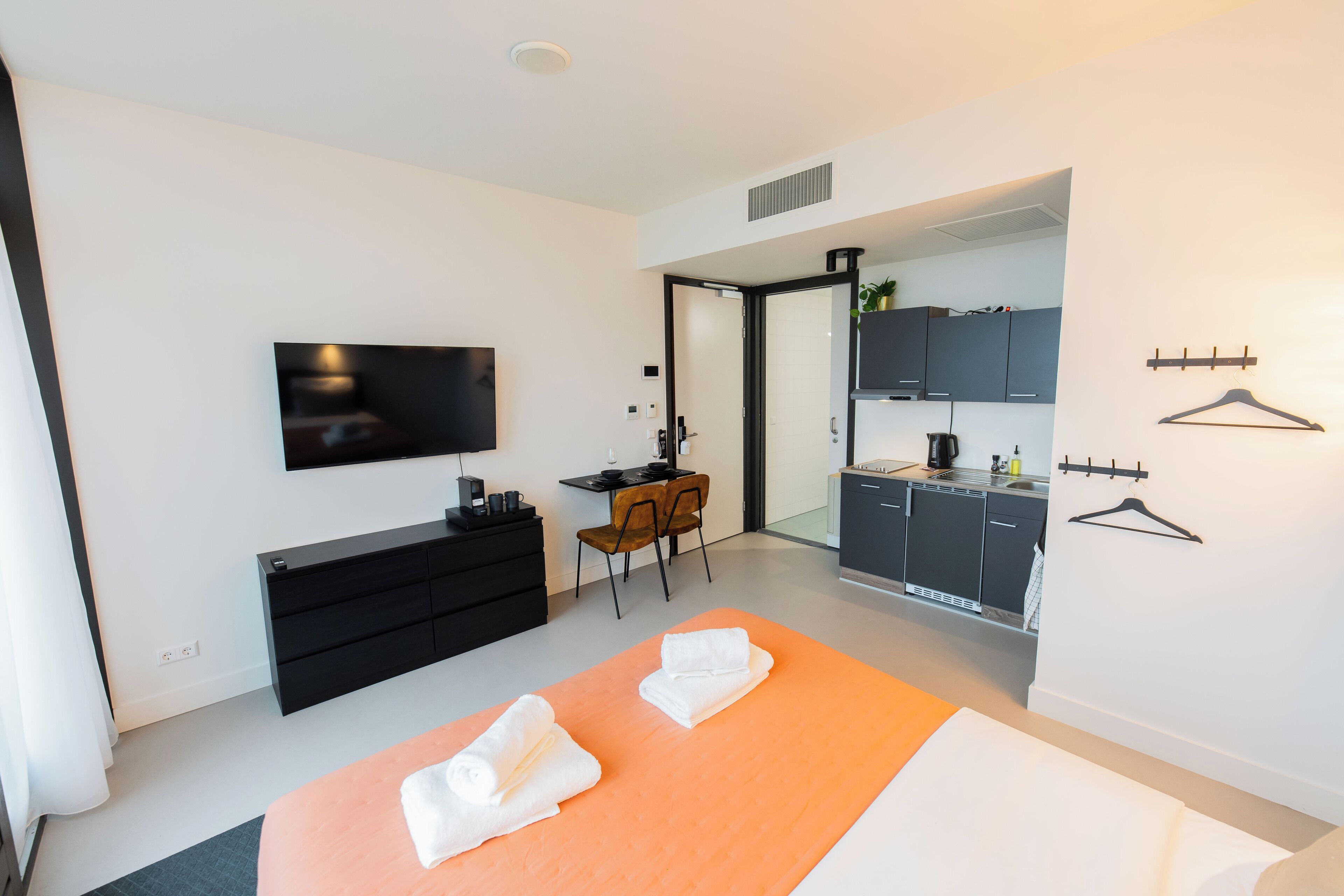 A modern hotel room with a bed, a desk, a TV, and a kitchenette.