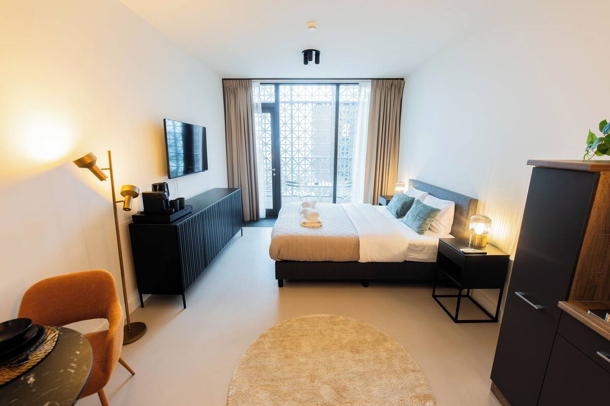 A modern hotel room with a bed, a desk, a chair, a TV, and a large window with curtains.
