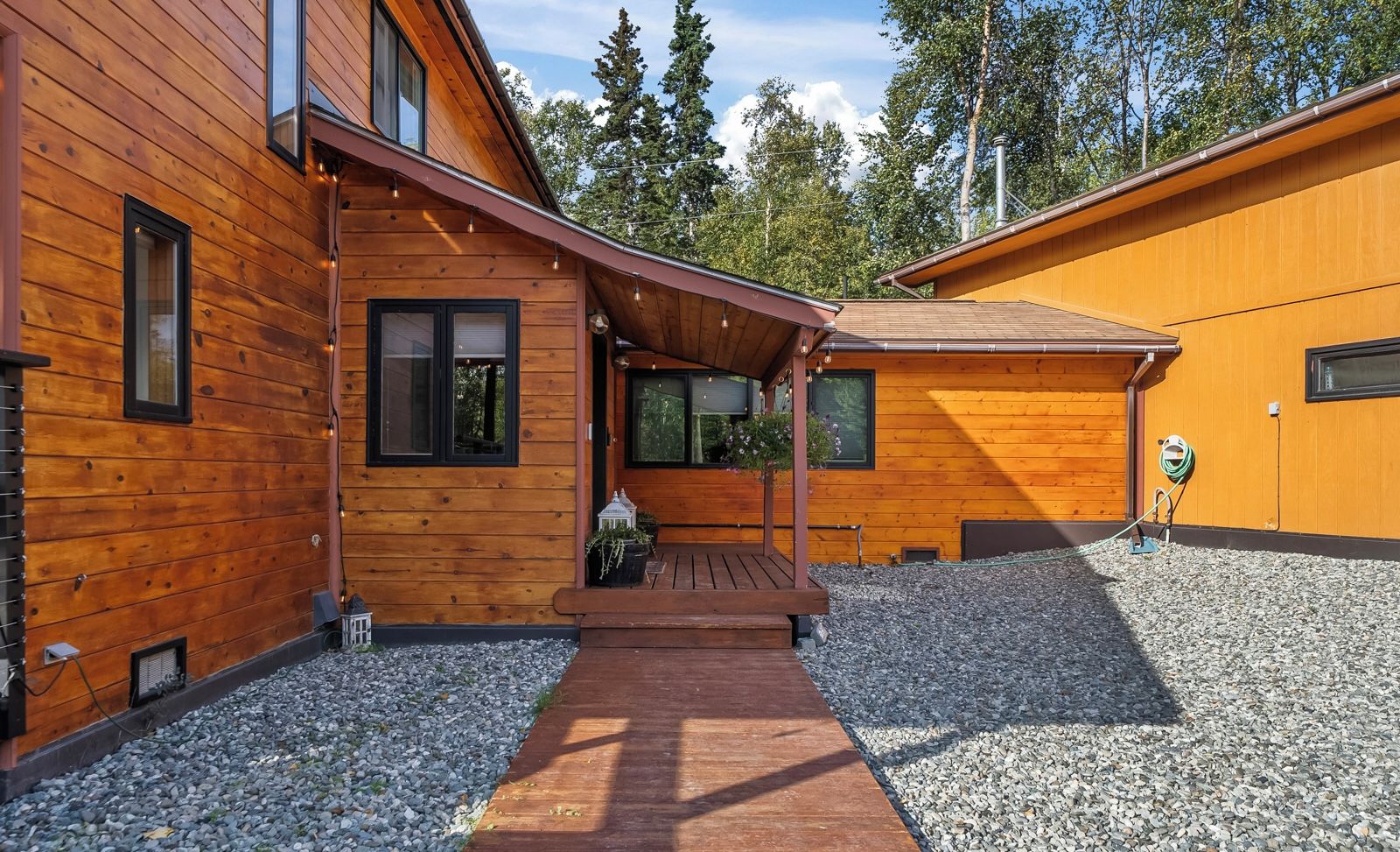 Una cabaña de madera con un porche cubierto, camino de grava y zona de jardín.