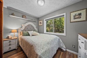 A bedroom with a bed, a nightstand, a dresser, a window with a view of trees, and a shelf with decorative items. - COZY HILLSIDE HOME W BEAUTIFUL DECK, FIRE PIT, SAUNA,BACKYARD & HUGE GAME-ROOM!! (Anchorage)