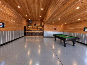 A pool table in a room with wooden walls and ceiling, a bar area with stools, and a star decoration on the wall. - COZY HILLSIDE HOME W BEAUTIFUL DECK, FIRE PIT, SAUNA,BACKYARD & HUGE GAME-ROOM!! (Anchorage)