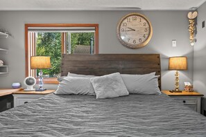 A bedroom with a bed, two bedside tables, a wall clock, a window, and a view of greenery outside. - COZY HILLSIDE HOME W BEAUTIFUL DECK, FIRE PIT, SAUNA,BACKYARD & HUGE GAME-ROOM!! (Anchorage)