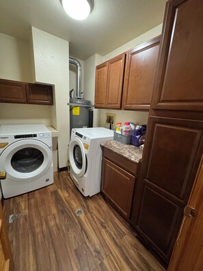 A compact laundry room with a front-loading washer and dryer, wooden cabinets, and a countertop with cleaning supplies. - COZY HILLSIDE HOME W BEAUTIFUL DECK, FIRE PIT, SAUNA,BACKYARD & HUGE GAME-ROOM!! (Anchorage)