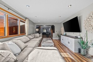 A modern living room with a large sofa, a flat-screen TV, and a wooden floor. - COZY HILLSIDE HOME W BEAUTIFUL DECK, FIRE PIT, SAUNA,BACKYARD & HUGE GAME-ROOM!! (Anchorage)