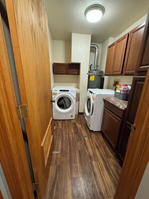 A laundry room with two washing machines, wooden cabinets, and a countertop with cleaning supplies. - COZY HILLSIDE HOME W BEAUTIFUL DECK, FIRE PIT, SAUNA,BACKYARD & HUGE GAME-ROOM!! (Anchorage)