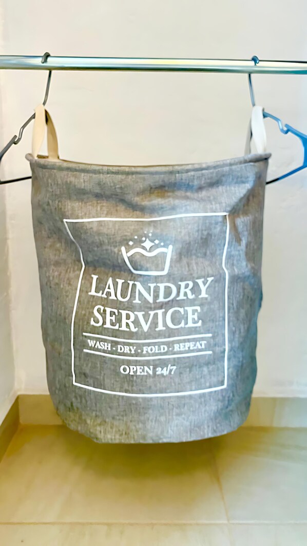 Una bolsa de lavandería gris colgada en un tendedero metálico, con un letrero que dice "Laundry Service" y "Abierto las 24 horas".