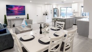 A modern kitchen with white cabinets, a large island, and a dining area with white chairs and a table set for two. - Newly Remodeled 2/2 Home with Golf Access, Game Room & Salt-Water Heated Pool (Cape Coral)