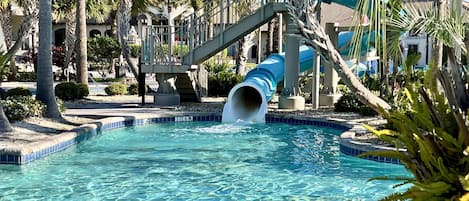 A pool with a water slide, palm trees, and a building with a balcony.