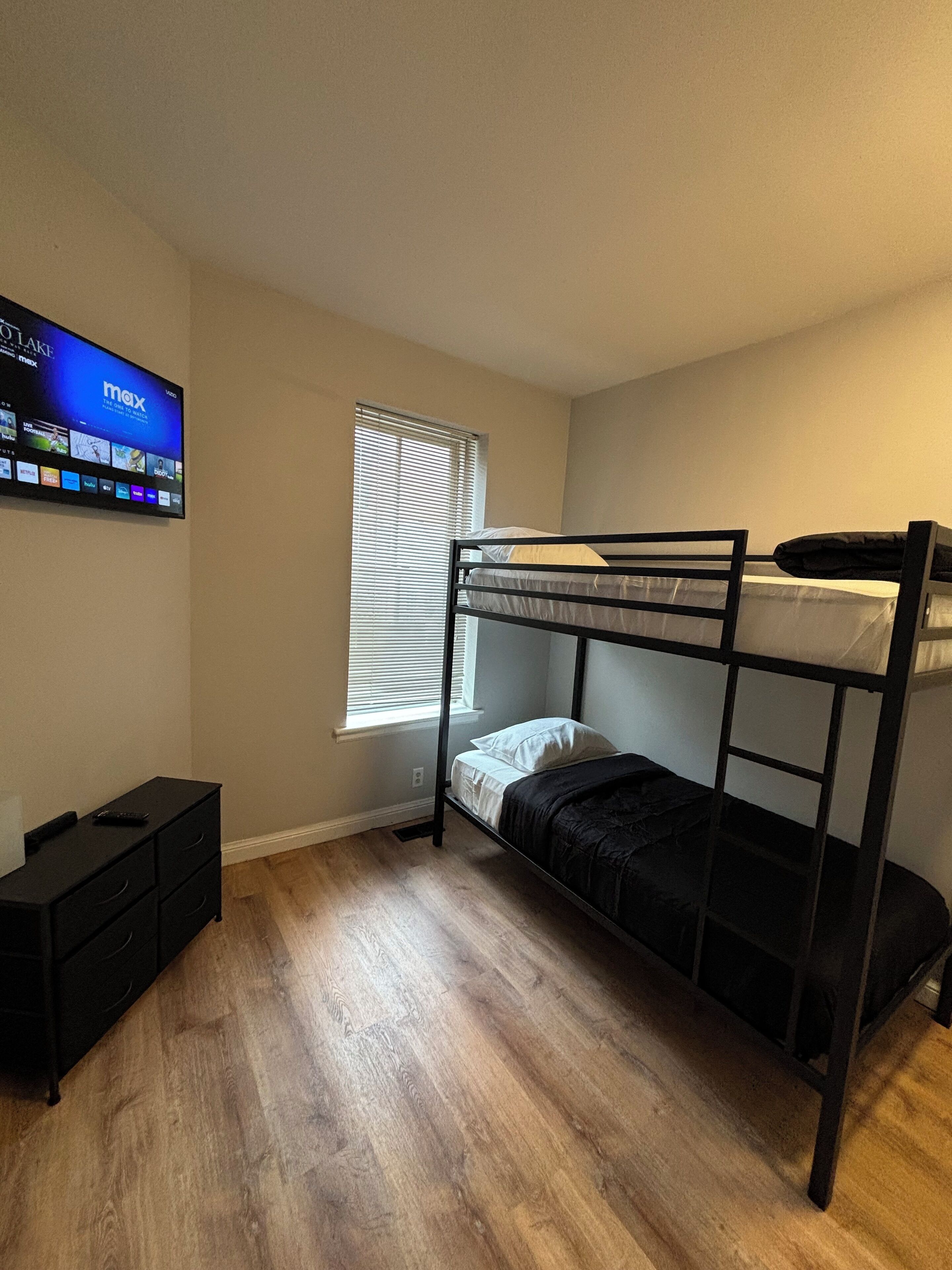 A room with a bunk bed, a flat-screen TV mounted on the wall, a small dresser, and a window with blinds.