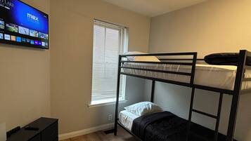 A room with a bunk bed, a flat-screen TV mounted on the wall, a small dresser, and a window with blinds.