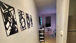 A hallway with a wooden floor, a gray rug, and black wall art. There is a kitchen area visible in the background with a refrigerator and a TV. - Cozy Soulard Getaway Near Downtown St. Louis (St. Louis)
