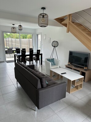 A modern living room with a sofa, TV, dining area, and a staircase. - Large house - Sleeps 8 - Jacuzzi - near St. Malo - 5 min. from the beach (Saint-Méloir-des-Ondes)