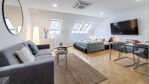 Ein moderner, gut beleuchteter Raum mit einem grauen Sofa, einem Bett mit grüner Decke, einem Esstisch mit Stühlen, einem Flachbildfernseher und Dachfenstern.