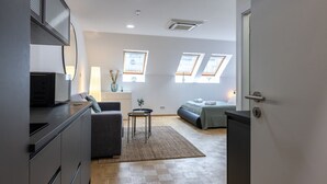 Ein moderner, kompakter Wohnraum mit Küchenzeile, einer Couch, einem kleinen Tisch und einem Bett. Dachfenster sorgen für natürliches Licht.