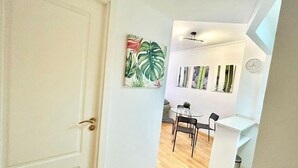 A hallway leading to a dining area with a white door, a wall with abstract art, a clock, and a small table with chairs. - Valleca Tren Sierra Guadalupe 1 (1 bedroom apartment) (Madrid)