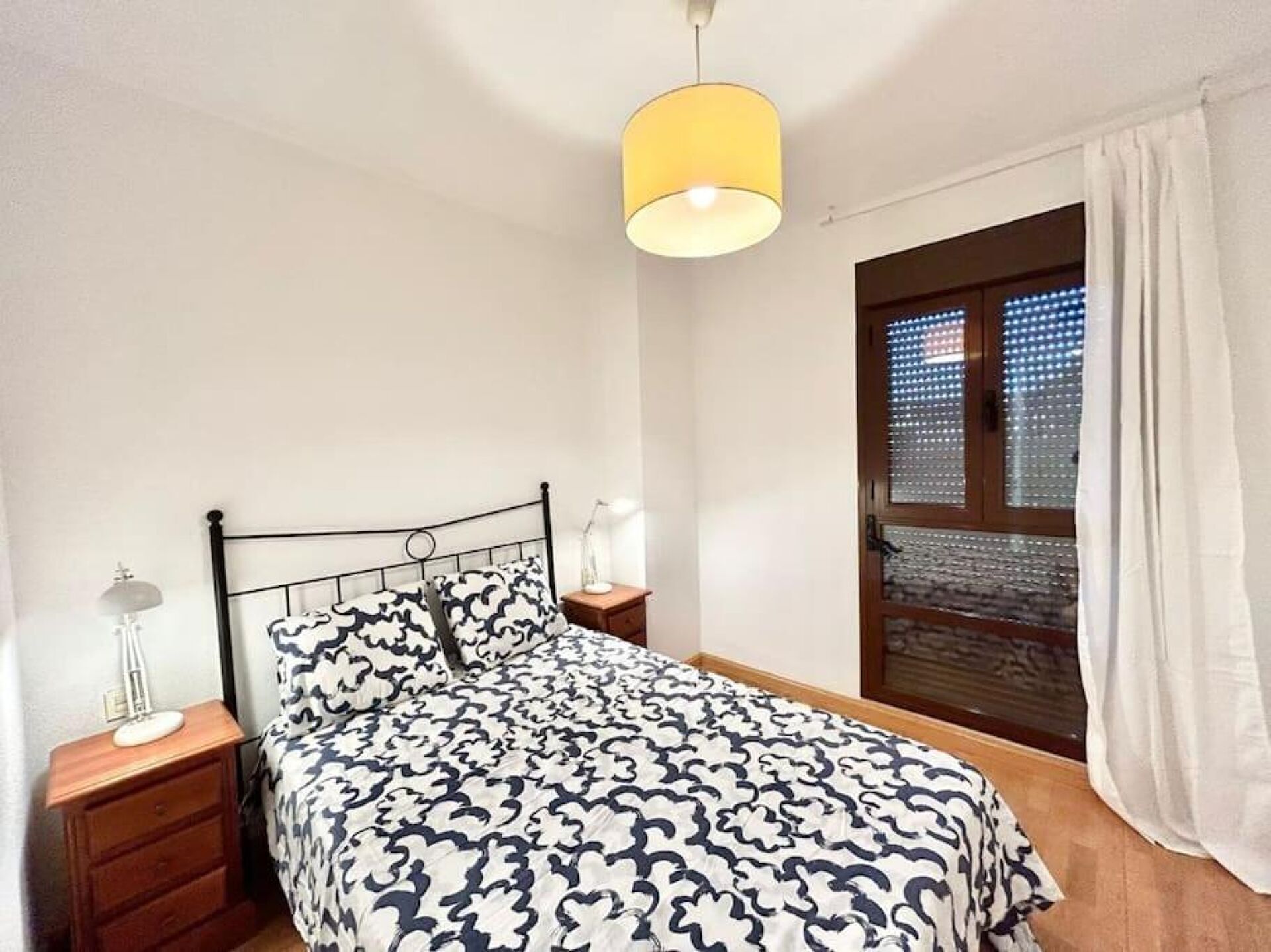 Un dormitorio con cama, mesitas de noche, lámpara, ventana con cortinas y una lámpara colgante.