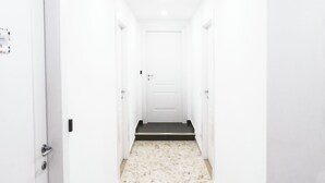 Un couloir étroit et blanc, avec une porte au fond, une petite marche menant à un plancher sombre, et un interrupteur sur le mur.