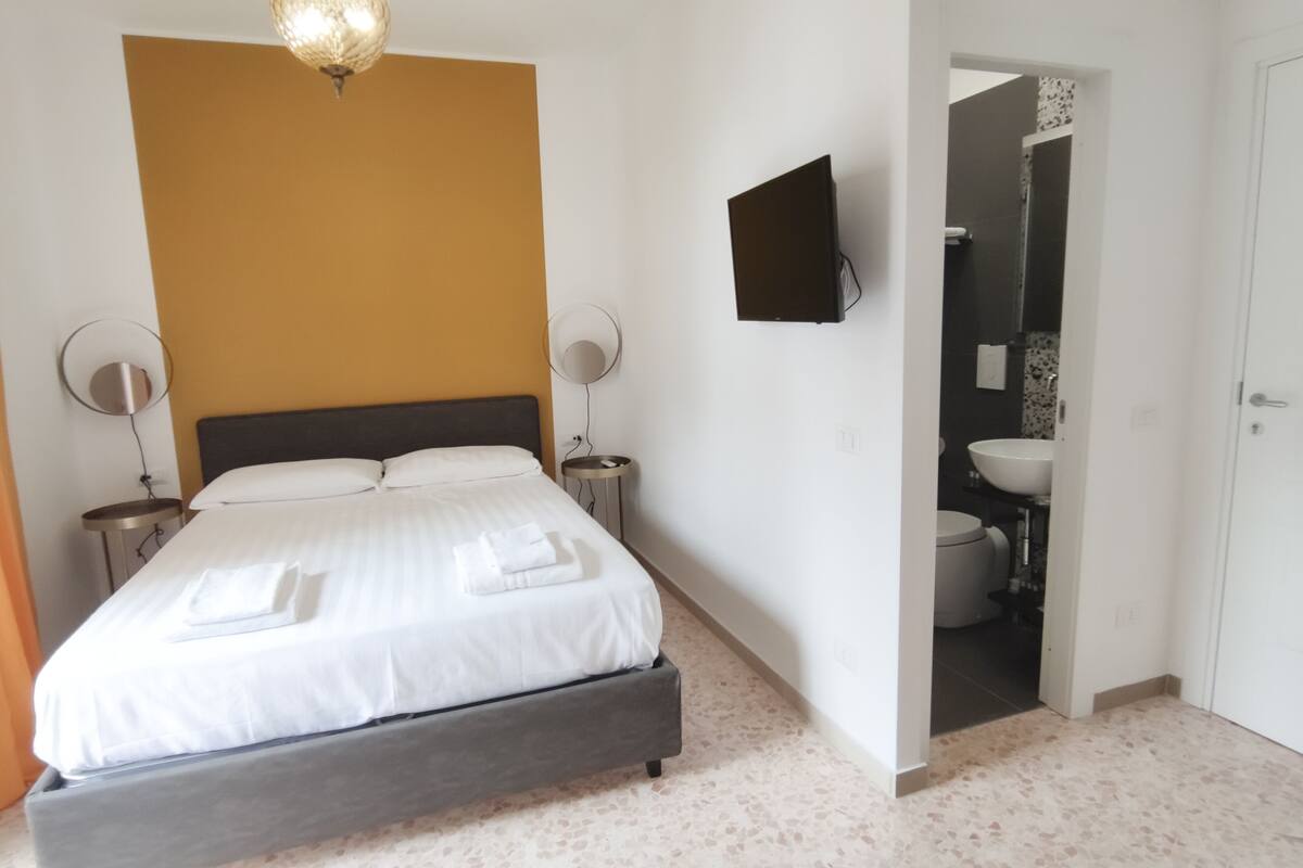 A bedroom with a bed, bedside tables, a wall-mounted TV, and a bathroom visible through an open door.