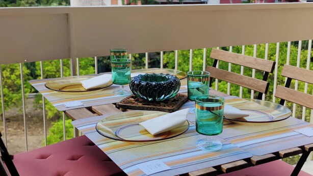 Un balcon avec une table dressée pour deux personnes, agrémentée d’un centre de table en verre, de deux verres et d’une serviette. Le balcon offre une vue sur une colline, avec des bâtiments et de la végétation.